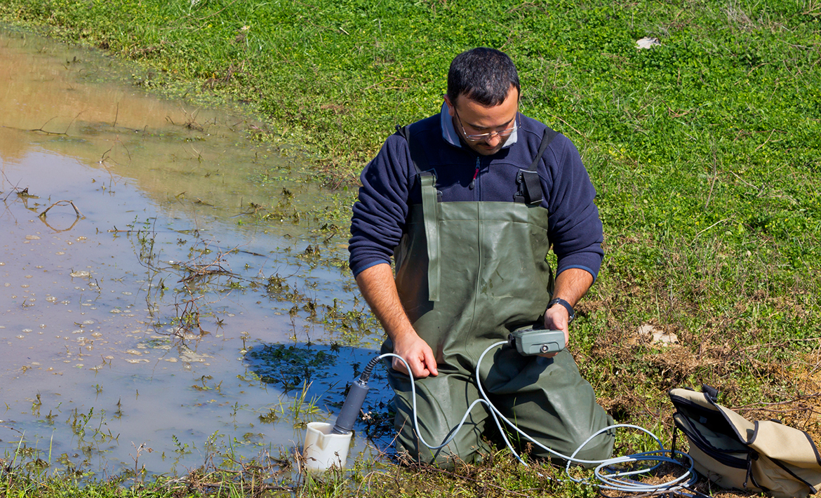 A man in overalls kneeling in grass. He is using an analytical instrument next to an area of water.